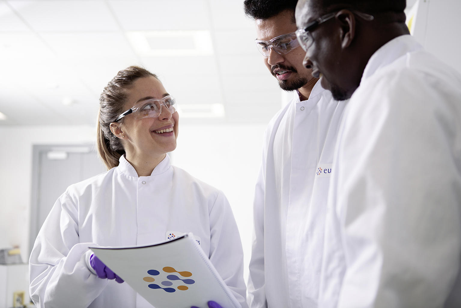 Three individuals in white lab coats are standing together in a laboratory setting. One person is holding a document with a colorful circular pattern on the cover. The individuals' faces are obscured for privacy