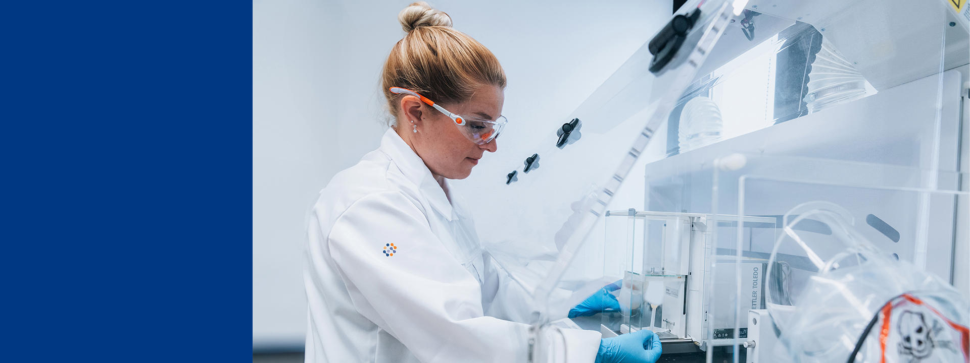 A person wearing a white lab coat and blue gloves is working in a laboratory, handling equipment inside a protective enclosure.