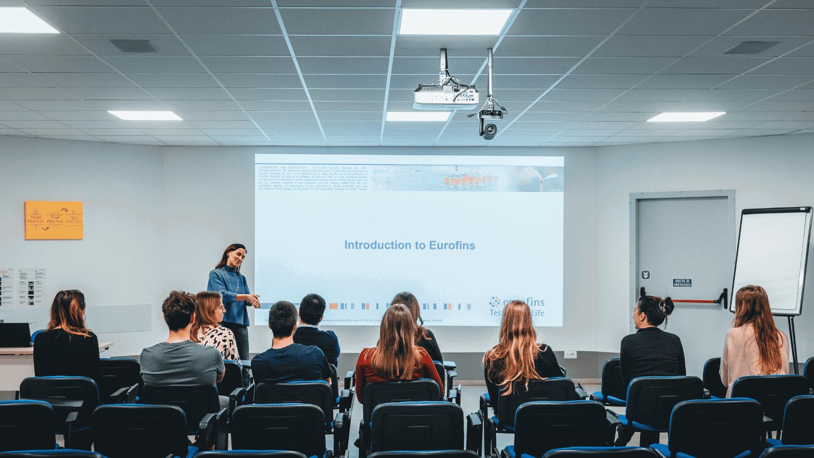 A classroom or conference room setting with people seated and facing a presenter standing in front of a large screen displaying the text 'Introduction to Eurofins.' Chairs are arranged in rows, and a ceiling-mounted projector and a whiteboard or flip chart are visible