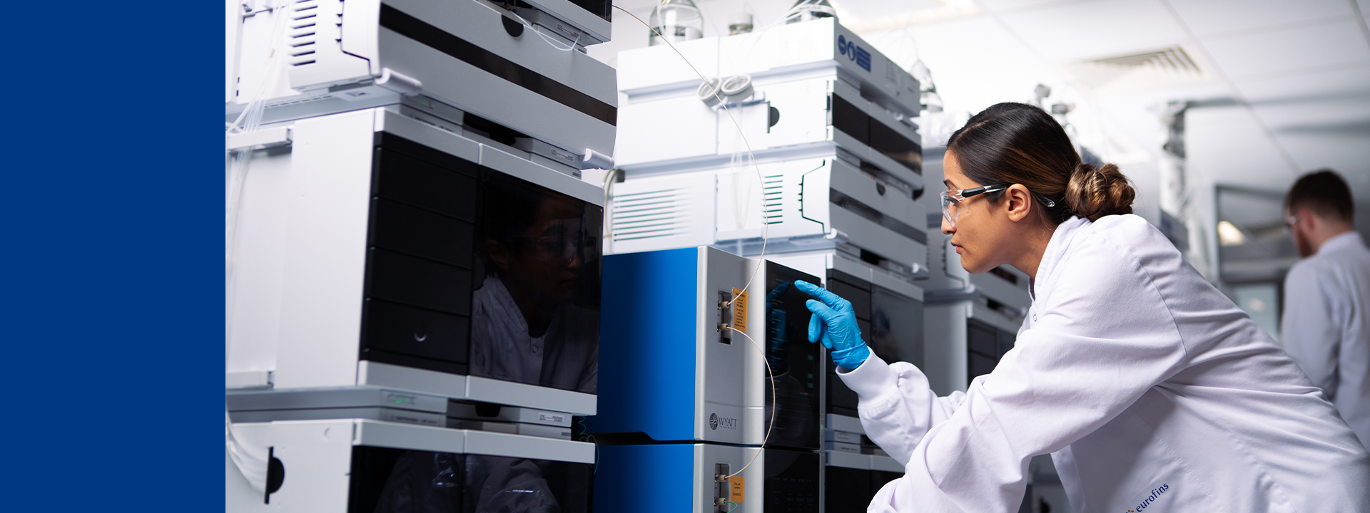 A laboratory technician wearing a white lab coat and blue gloves operates an ICE imaged capillary electrophoresis equipment in a modern laboratory. The technician is adjusting settings on the equipment, surrounded by other similar machines with various cables and tubes connected. Another person in the background is also working in the lab.