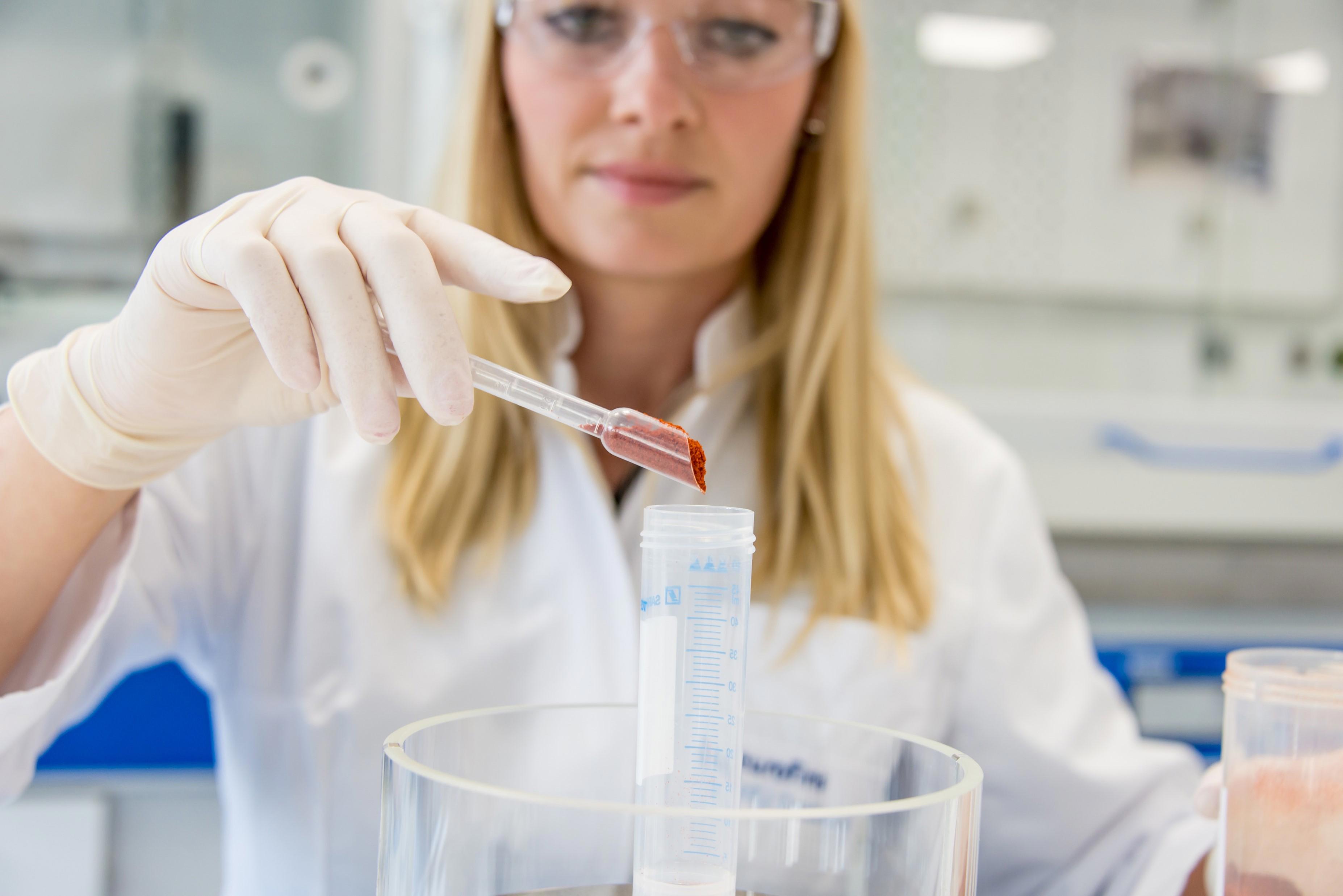 A person wearing a white lab coat and gloves is holding a pipette with a red powder, transferring it into a graduated cylinder inside a laboratory setting. The background shows laboratory equipment and shelves. The person's face is slightly out of focus.