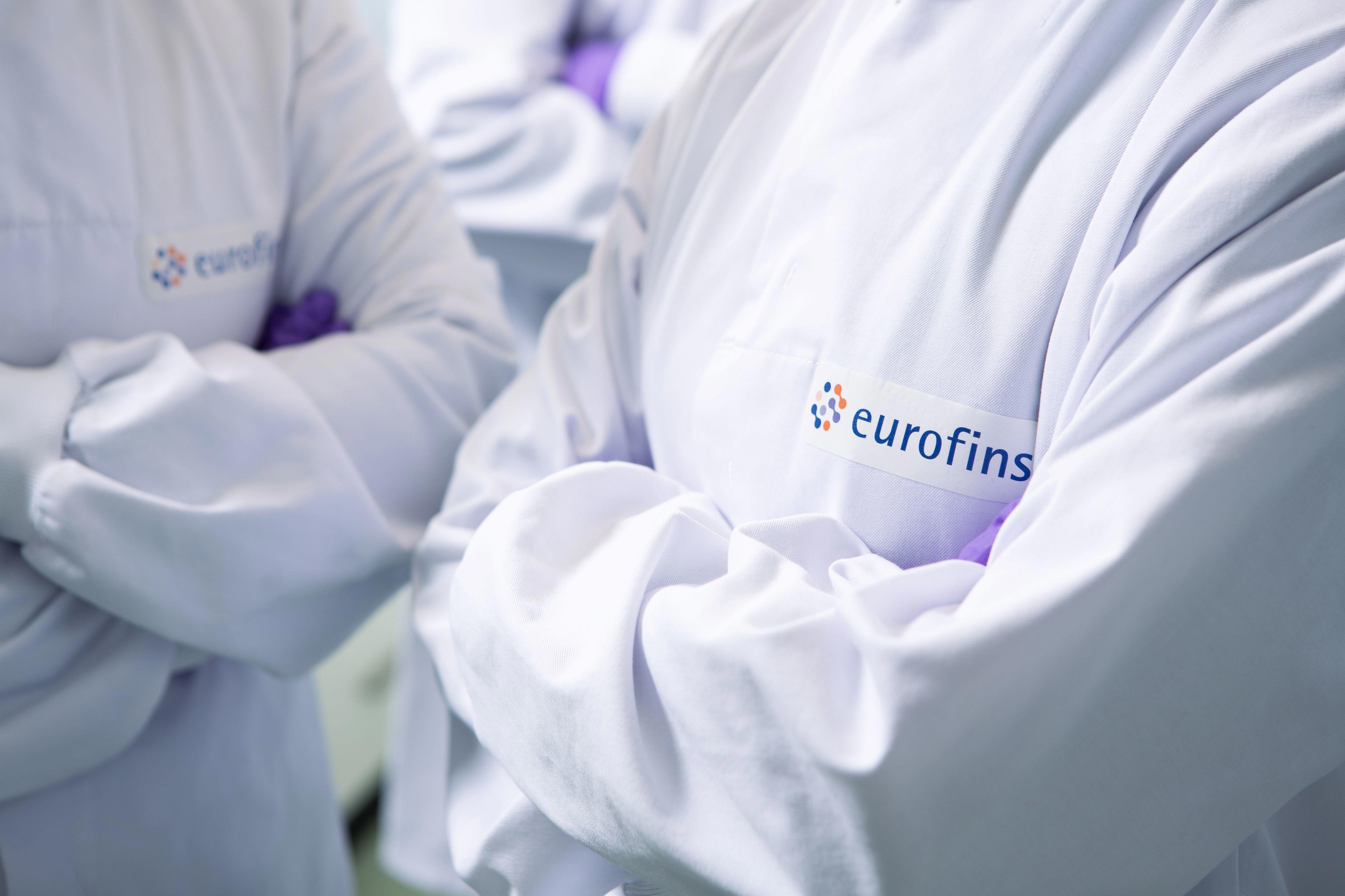 Image is zoomed in on two scientists who are wearing white lab coats displaying a eurofins logo with their arms crossed