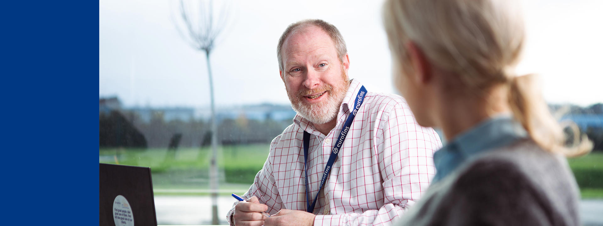 A man wearing a checkered shirt and a eurofins ID lanyard, is sitting at a table with a laptop in front of him. He is holding a pen and appears to be engaged in conversation with another person whose back is facing the camera. The background shows an outdoor scene with greenery and trees.