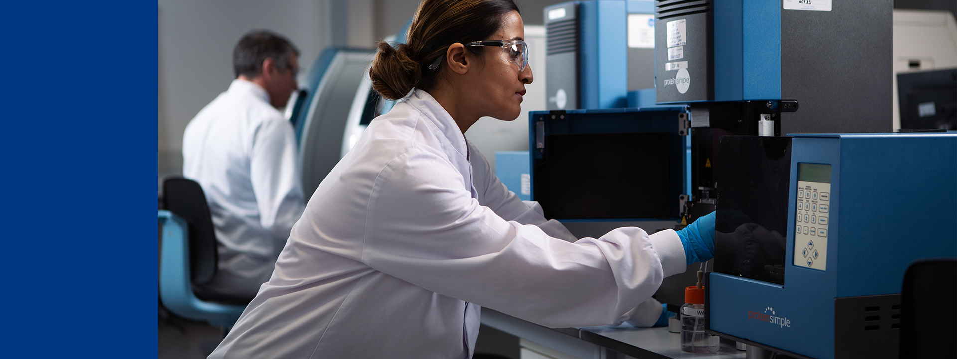 A laboratory technician wearing a white lab coat and blue gloves is operating a blue and black ICE imaged capillary electrophoresis instrument on a countertop. Another person in the background is also working with similar equipment.