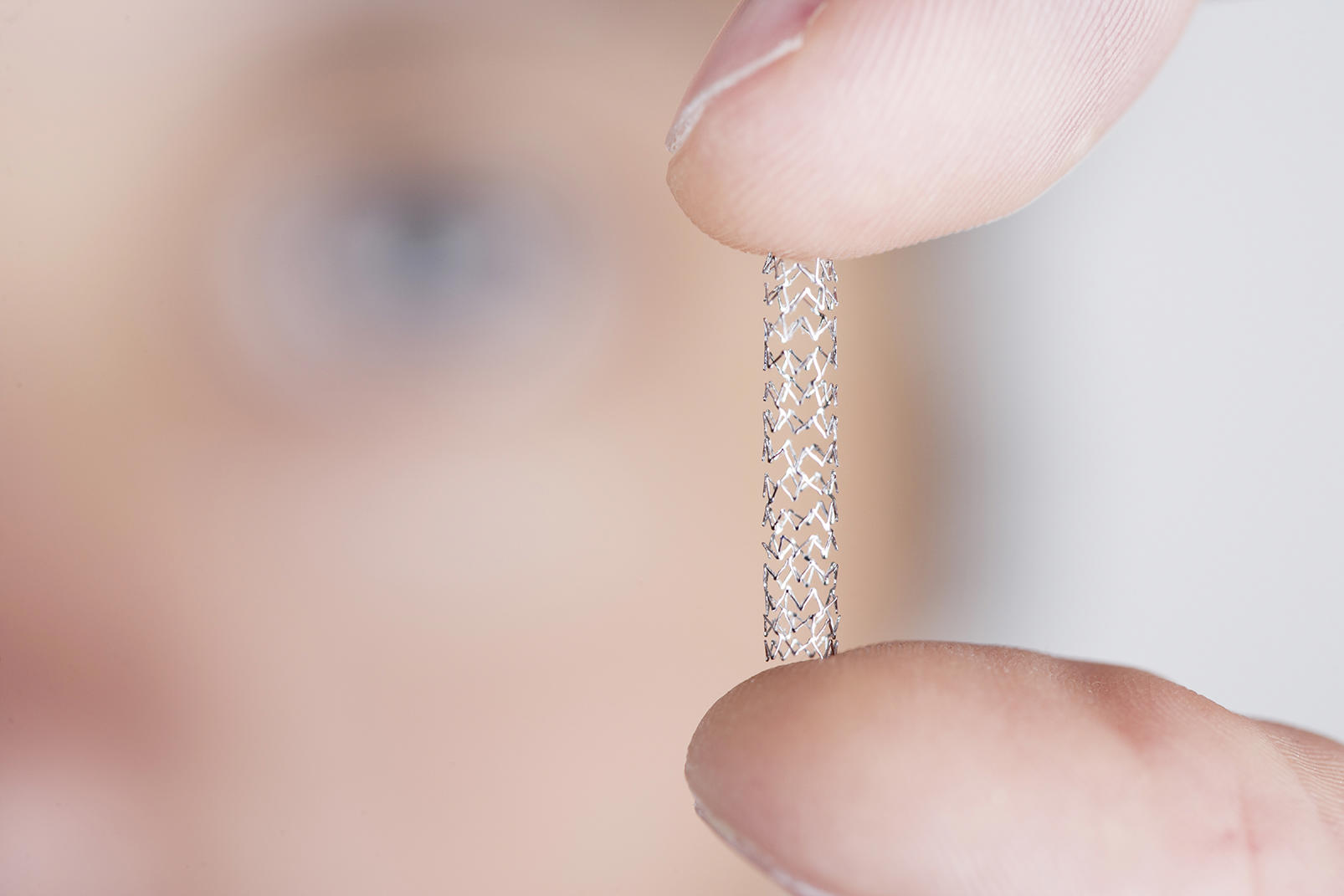 Close-up of a person's fingers holding a small, cylindrical, mesh-like medical device, likely a stent. The background is blurred, and the person's face is slightly visible but out of focus