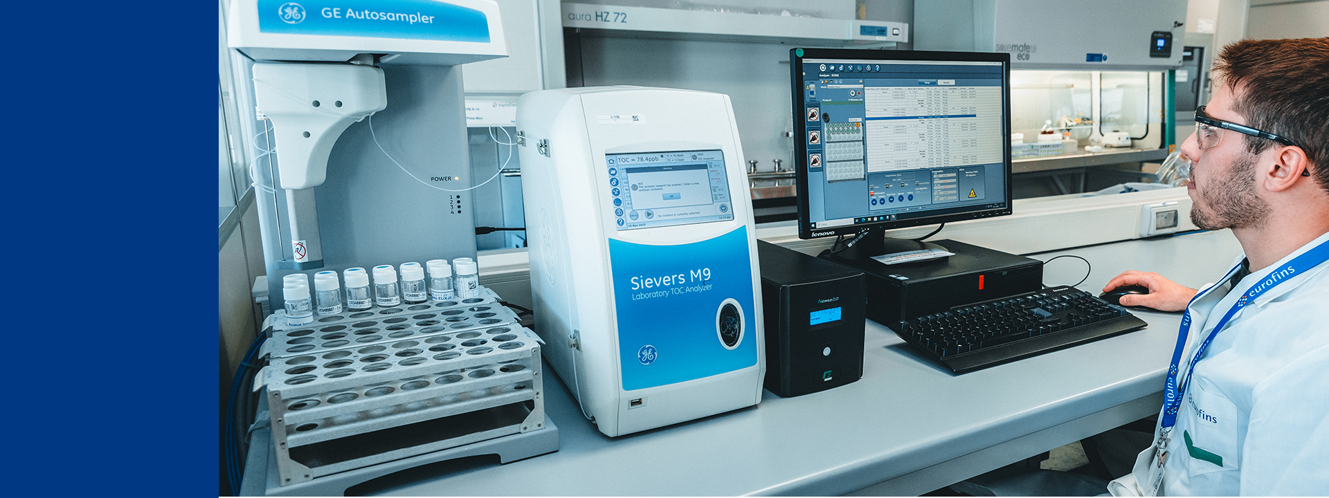 Laboratory setting with a technician in a lab coat working at a computer. The lab equipment includes an automated sample processor labeled 'GX-271 Liquid Handler' and another device labeled 'Seahorse XFp.' The technician is seated at the desk, interacting with the computer, which displays data and graphs. This environment is focused on the analysis and testing of excipients, bulkware, and active pharmaceutical ingredients (APIs), ensuring that these components meet the required quality standards.