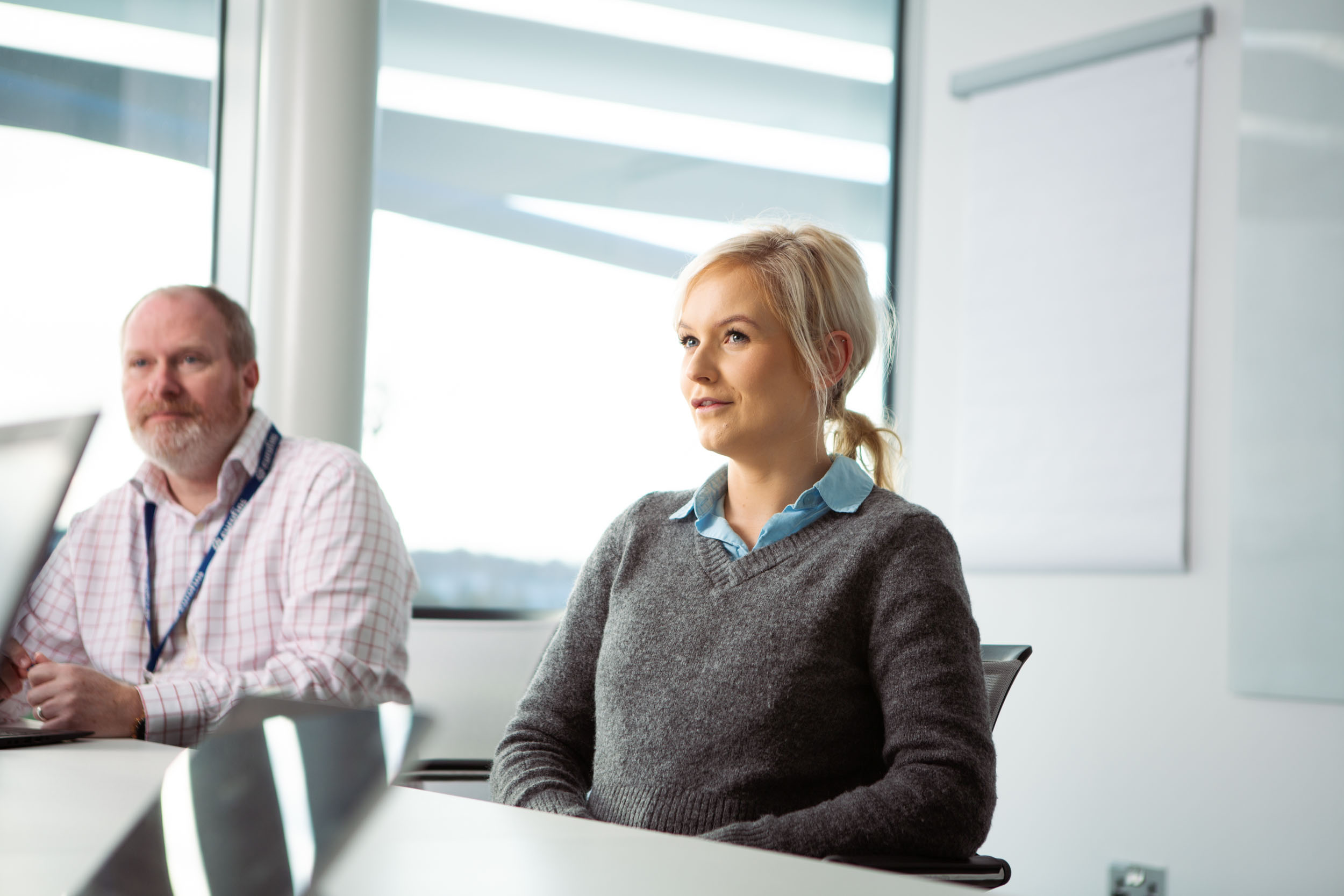 Two people sitting in an office or conference room. One wears a light-colored checkered shirt with a lanyard, and the other wears a grey sweater over a blue collared shirt. Large windows and a whiteboard or flip chart are in the background