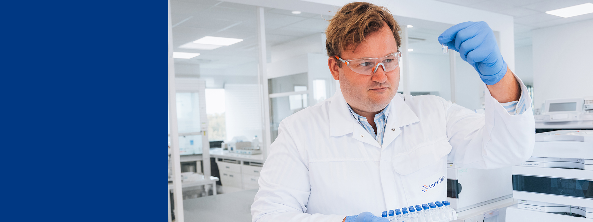 A scientist in a laboratory is holding a blue-capped test tube rack with multiple test tubes. The scientist is wearing a white lab coat with the logo "eurofins", blue gloves, and has short hair. The background shows laboratory equipment and workstations.