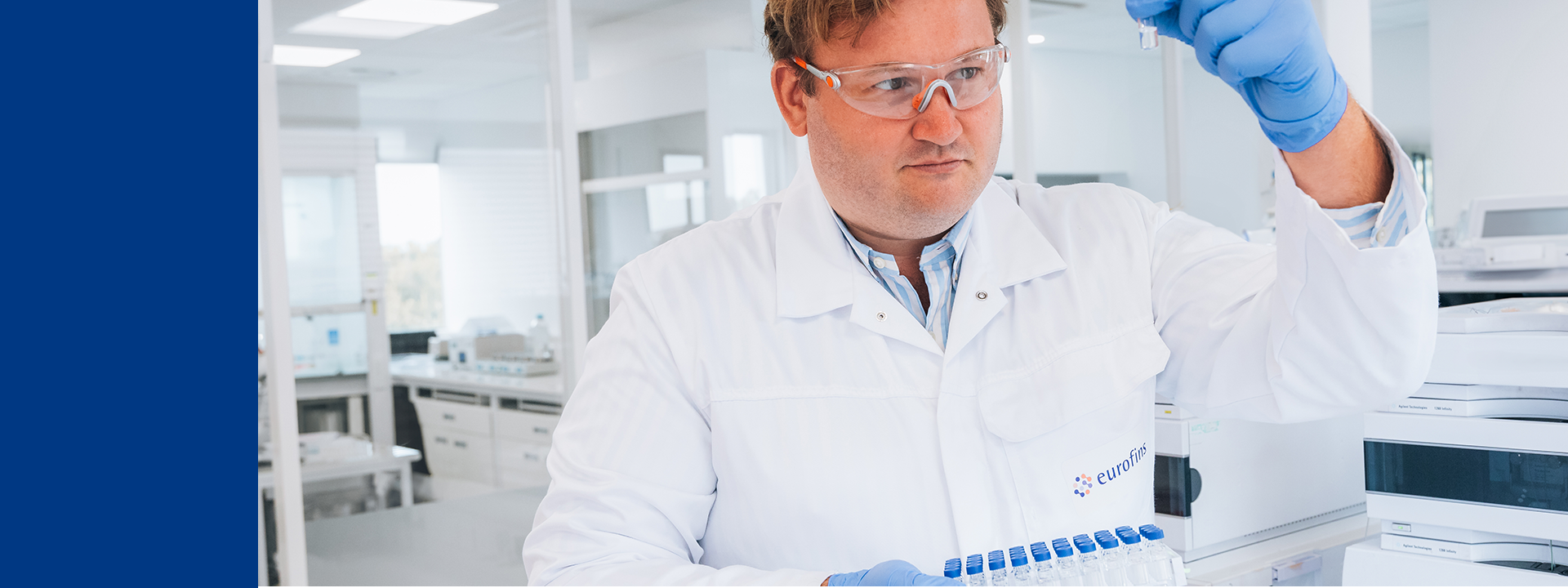 A scientist in a laboratory is holding a blue-capped test tube rack with multiple test tubes. The scientist is wearing a white lab coat with the logo "eurofins", blue gloves, and has short hair. The background shows laboratory equipment and workstations.