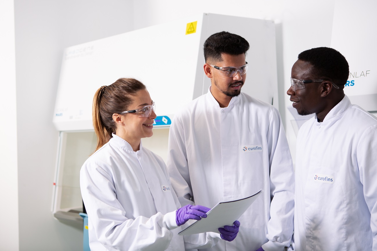 Team of scientists in lab coats engaged in discussion while reviewing documents in a microbiology lab, highlighting teamwork in the production and banking of microbial strains.