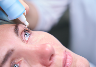 Close-up image of a person receiving eye drops. A blue eye drop bottle is held above the open eye, with a droplet about to fall. The person has blue eyes and is looking upward during the administration.