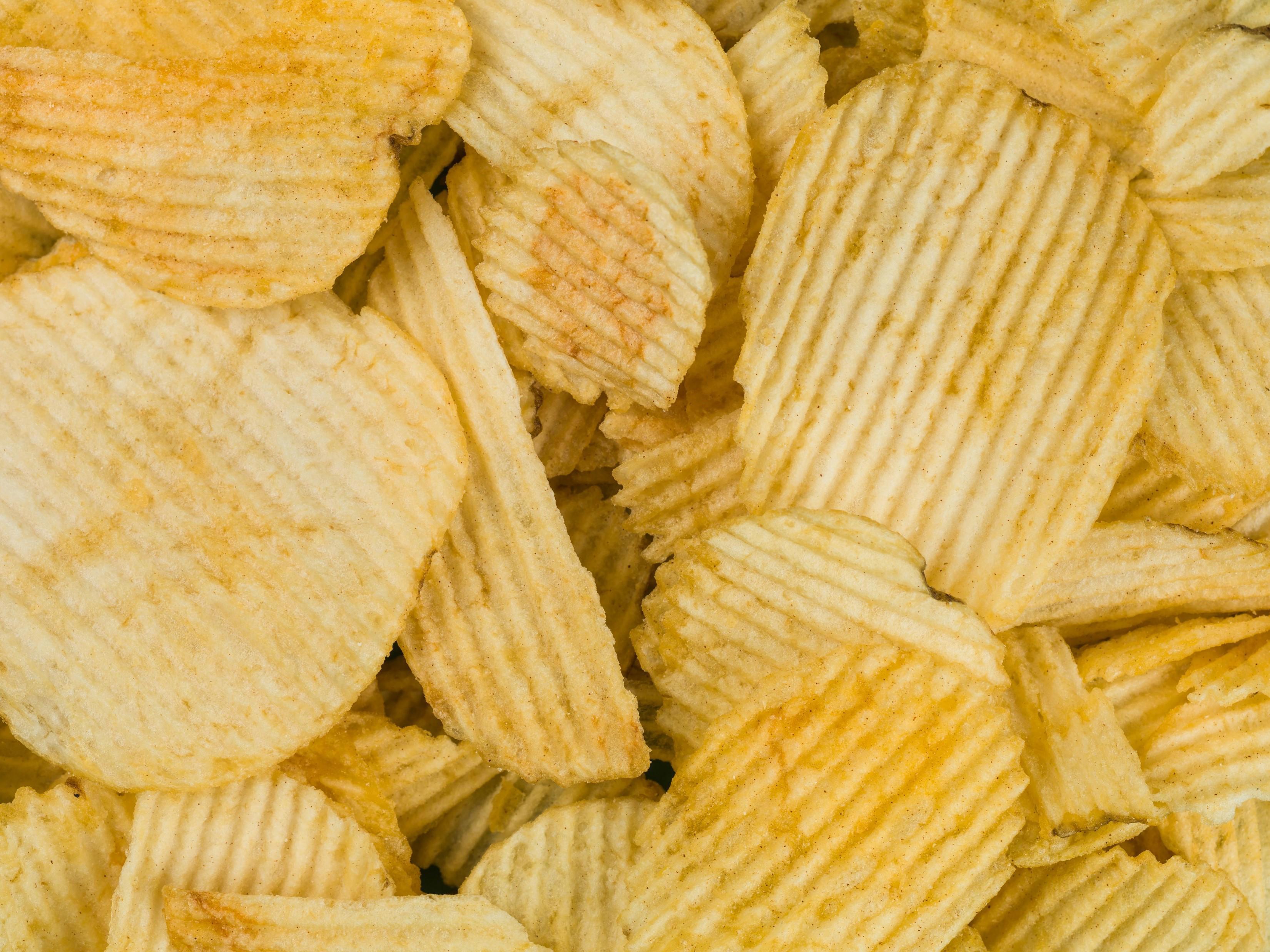 A close-up image of a pile of ridged potato chips. The chips are golden yellow in colour and have a textured, wavy surface. Some chips are whole while others are broken into smaller pieces.