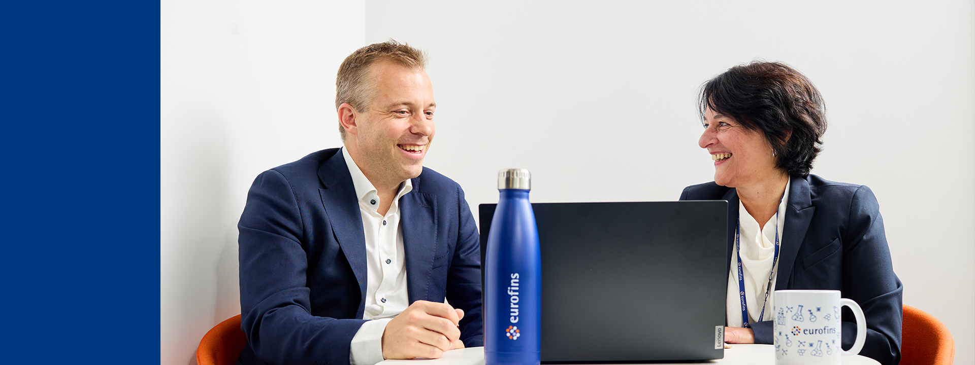 Two people in business attire are sitting at a round table with a laptop, a blue water bottle labeled 'eurofins,' and a white mug also labeled 'eurofins.'