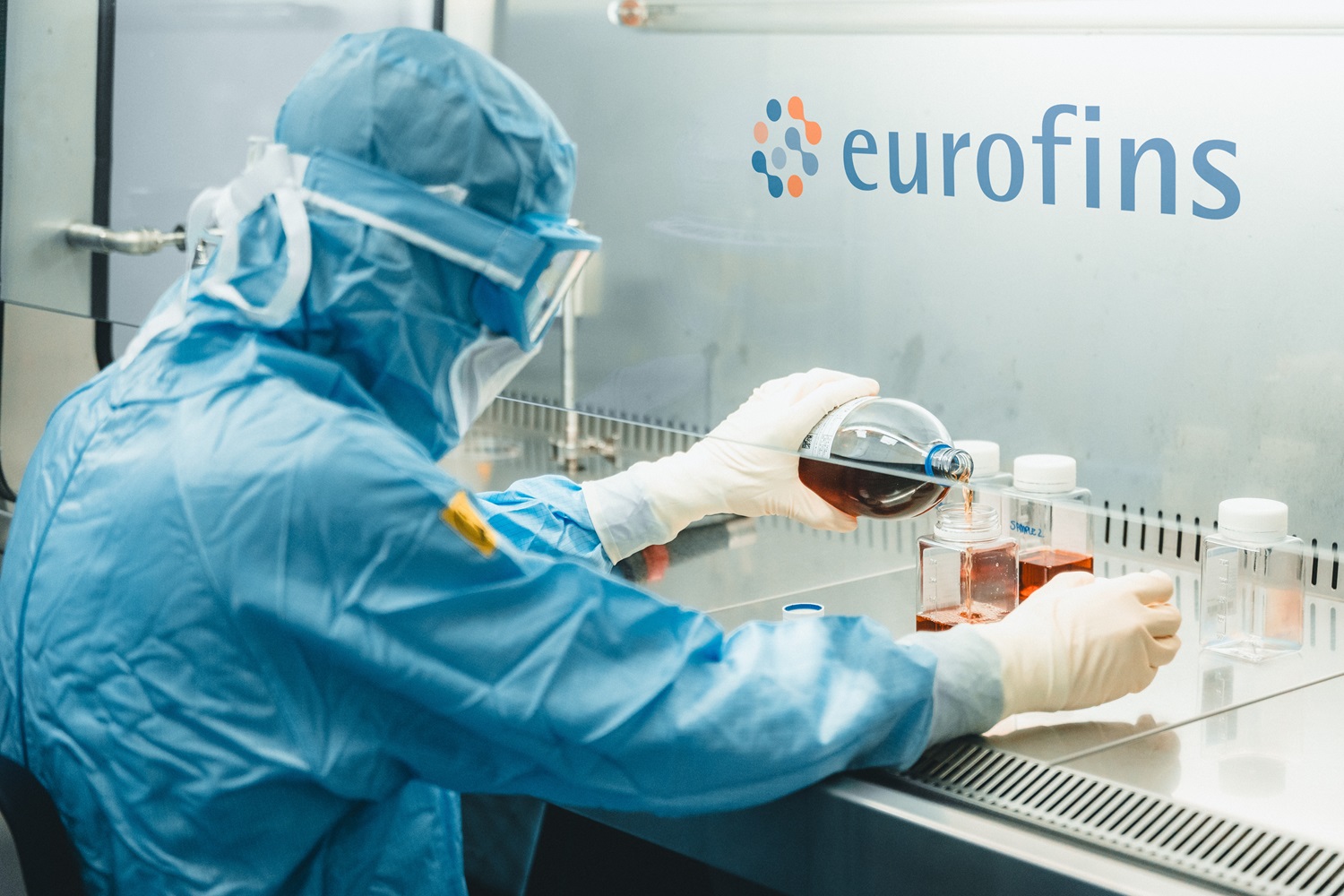 A scientist in full protective gear pours liquid between containers for sterility testing