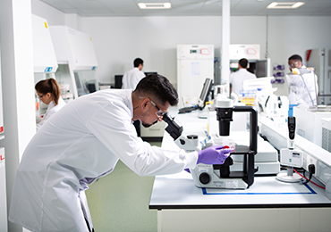 Scientists in a laboratory wearing white lab coats work with various instruments. One technician in the foreground uses a microscope, while others perform analytical tasks in the background. The setting reflects advanced testing procedures for empty and full capsid characterisation.