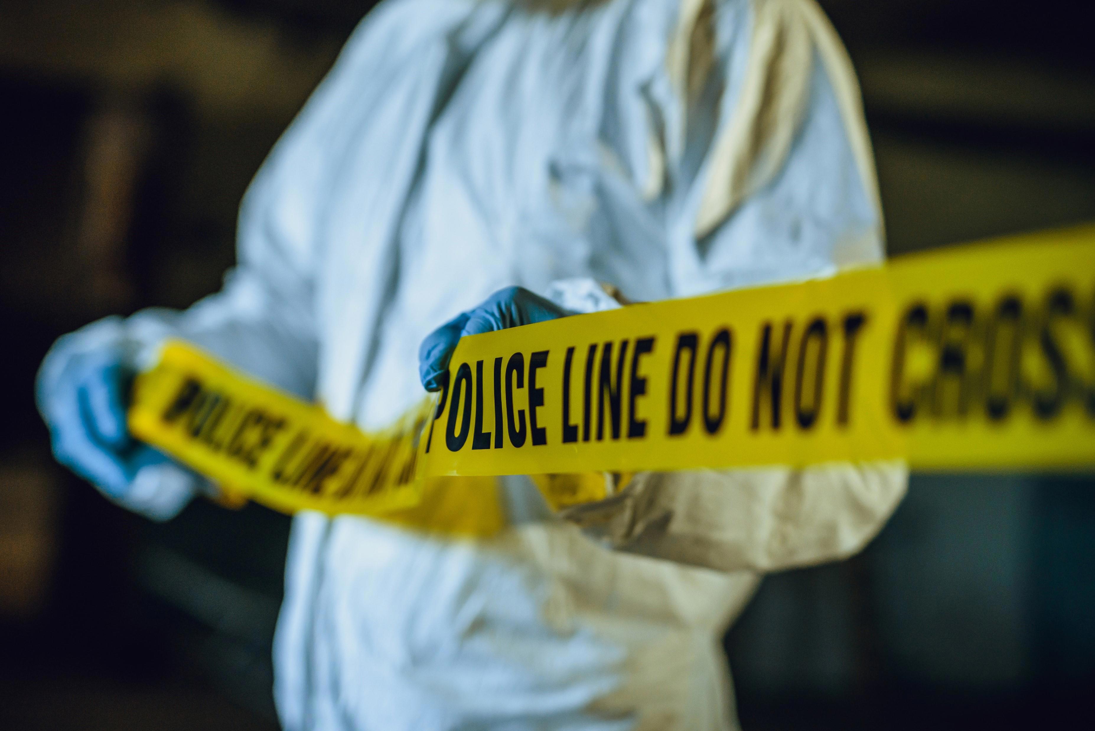 A person wearing a white protective suit and blue gloves is holding yellow police tape that reads "POLICE LINE DO NOT CROSS." The background is dark and out of focus.