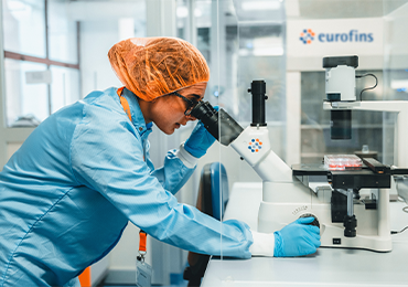 A laboratory technician in a blue lab coat, orange hairnet, and blue gloves examines samples under a microscope branded with the Eurofins logo. The setting highlights analytical procedures used in the characterisation of large molecules.