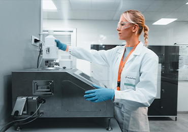 A person in a laboratory setting wearing a white lab coat, blue gloves, and an orange lanyard, operating or adjusting a piece of laboratory equipment. The background includes additional lab equipment and cabinets. The image is labeled 'Combination Product'.