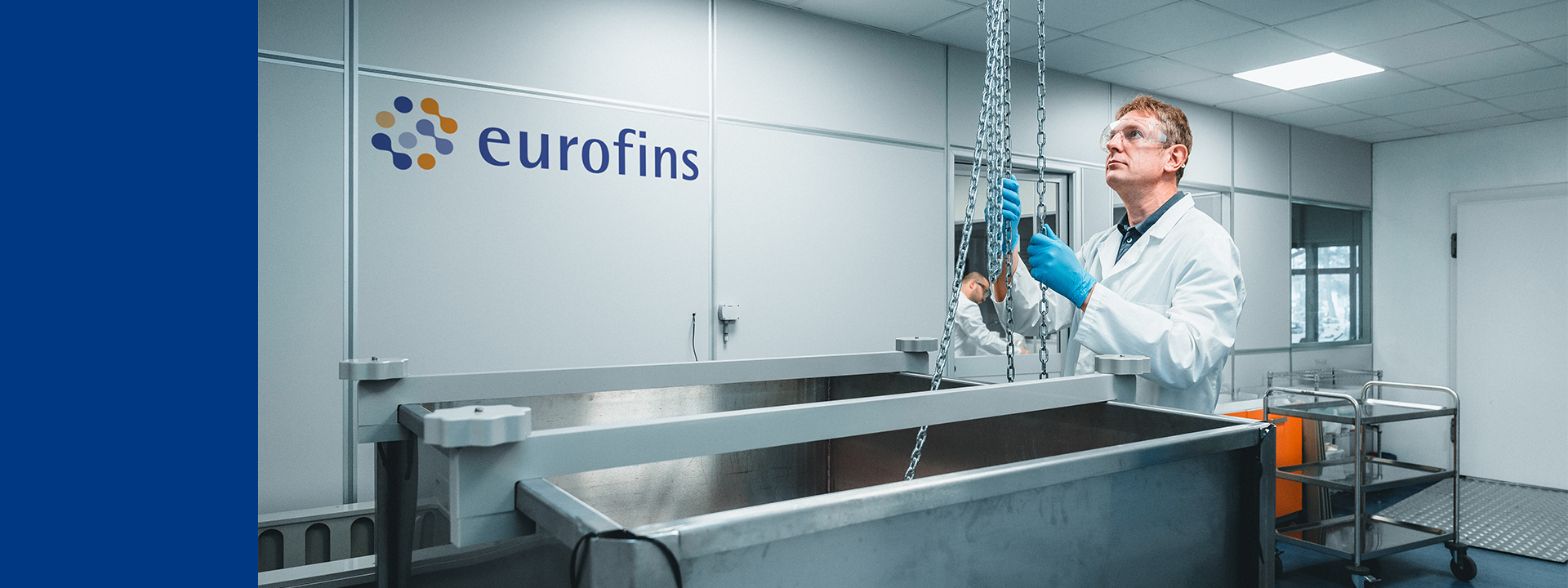 Laboratory setting with a technician wearing blue gloves and a white lab coat, working with equipment. The technician is handling chains connected to an overhead hoist, which appears to be lifting or lowering a large metal container. This environment is focused on container closure integrity tests, which involve ensuring that pharmaceutical containers are properly sealed to prevent contamination and maintain the sterility and stability of the product.