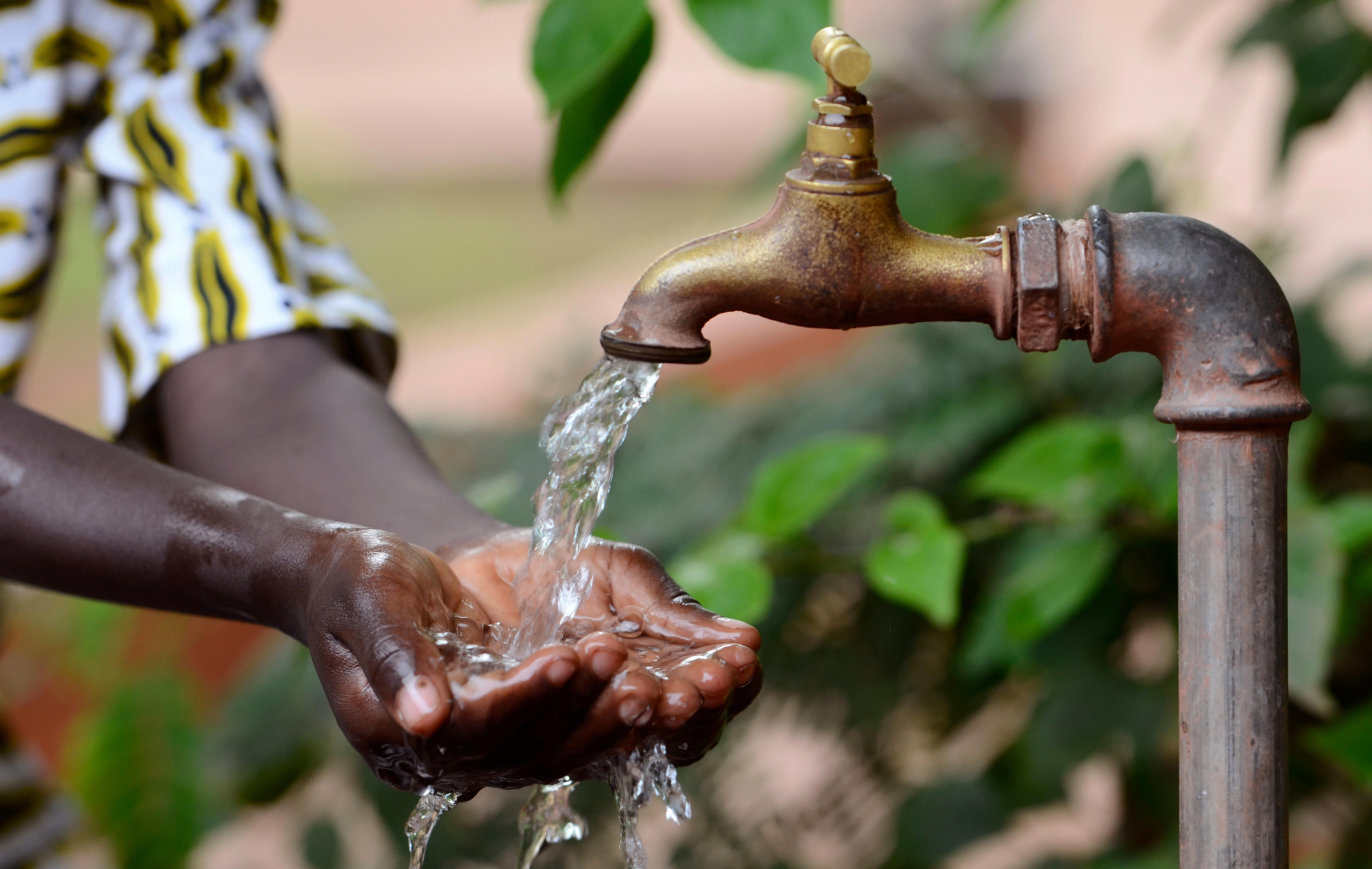 A close-up image of a person's hands being washed under a running outdoor faucet. The faucet is made of metal and has a brass handle. Water is flowing from the faucet into the person's cupped hands. The background shows green leaves and blurred natural surroundings, indicating an outdoor setting.