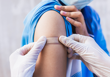 Close-up image of a person's upper arm with a patch being applied by a nurse wearing white gloves. The person is wearing a blue shirt and has lifted the sleeve to expose the application area.