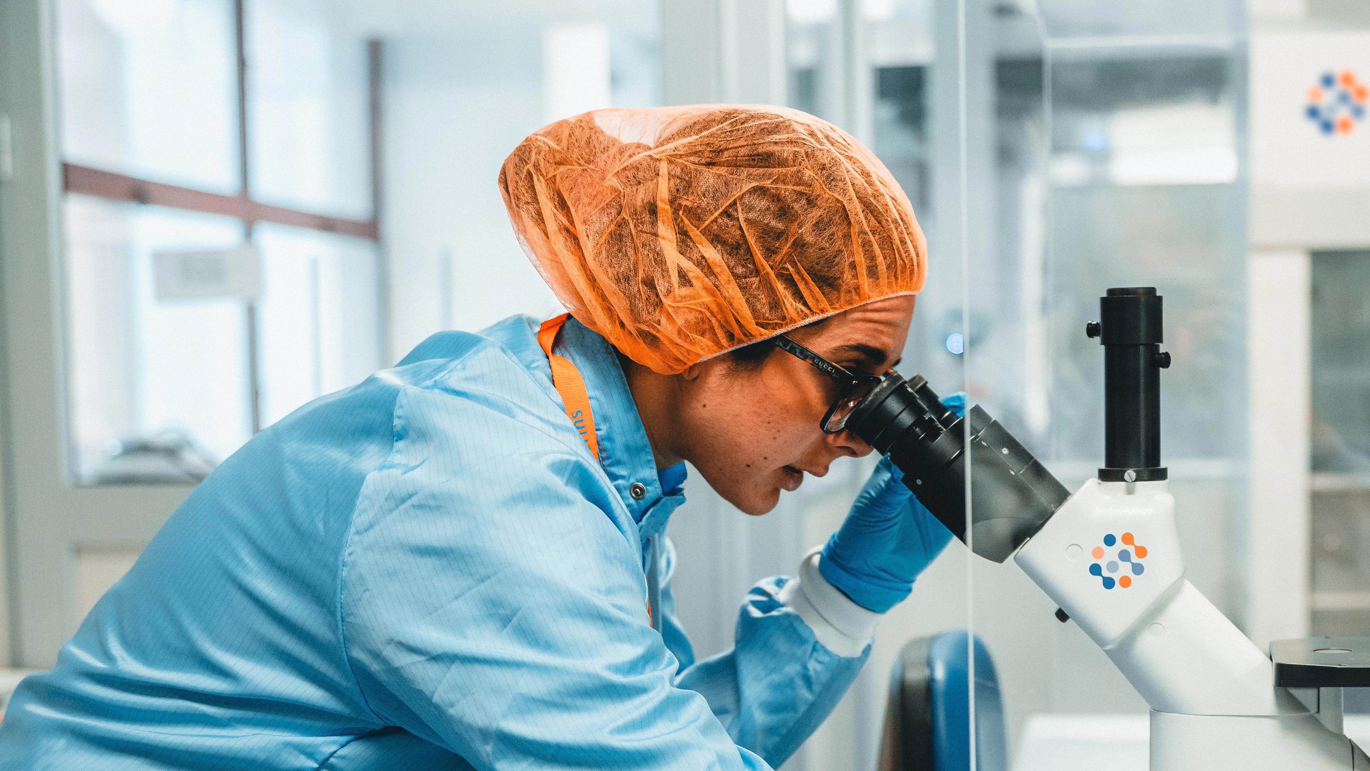 A person in a laboratory setting using a microscope. The person is wearing protective clothing, including a blue lab coat, gloves, and an orange hairnet. The microscope is positioned on a white table with various other laboratory equipment visible in the background. The Eurofins icon is on the microscope