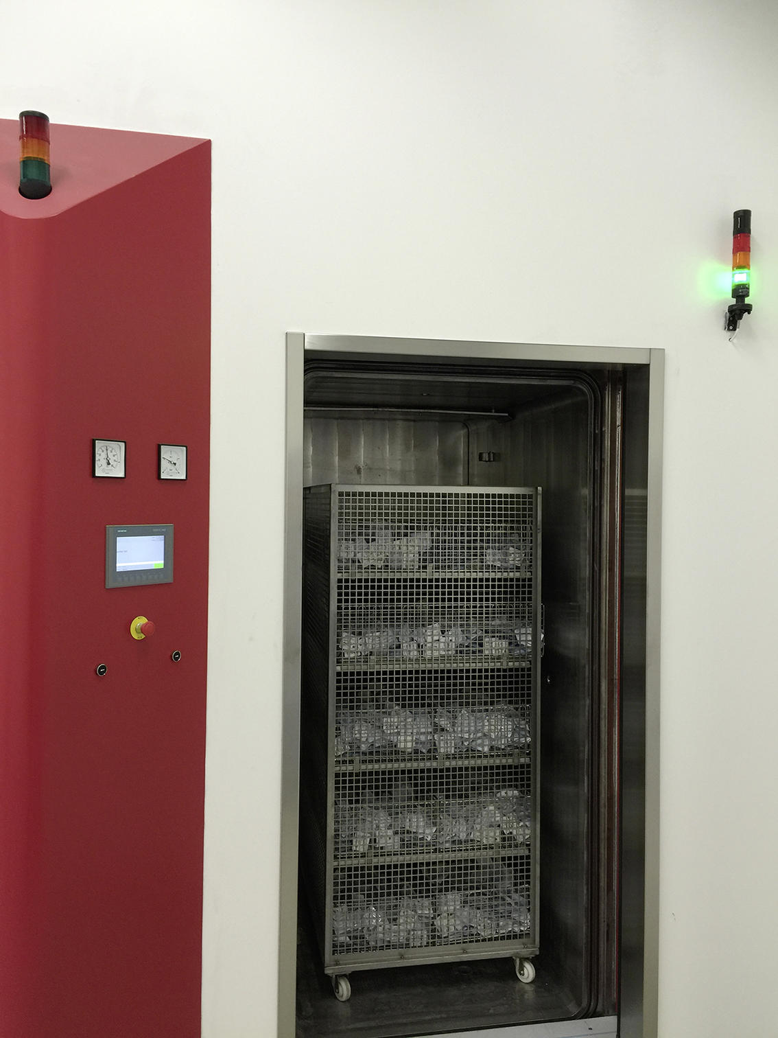 Sterilisation chamber with an open door, showing a metal rack with multiple shelves containing items in sterilisation packaging. A red control panel with two analogue gauges, a digital display, and an emergency stop button is visible on the left.